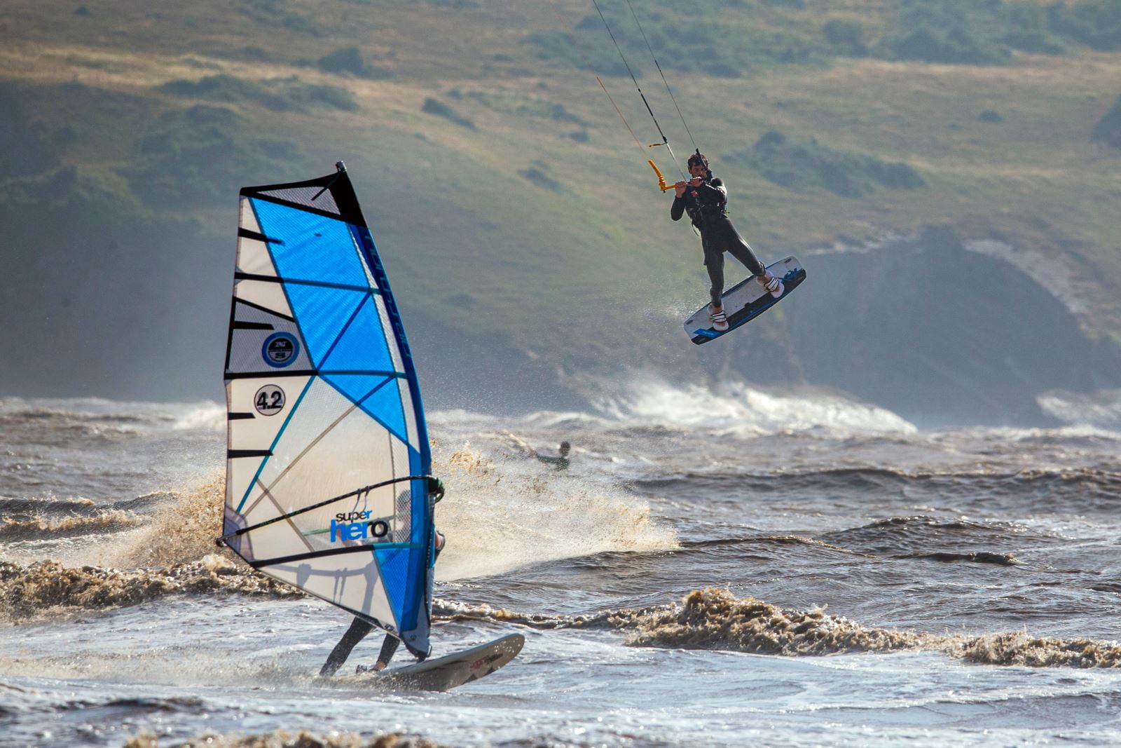 A kite surfer rides above a windsurfer